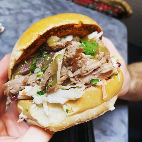 Close-up of a pulled pork sandwich with coleslaw and green sauce on a brioche bun.