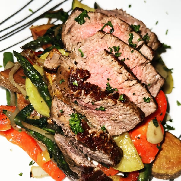 Sliced steak with a pink center, drizzled with sauce, served over roasted vegetables and potatoes.