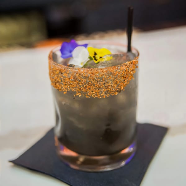 Dark cocktail with a spiced rim, garnished with purple, white, and yellow edible flowers.